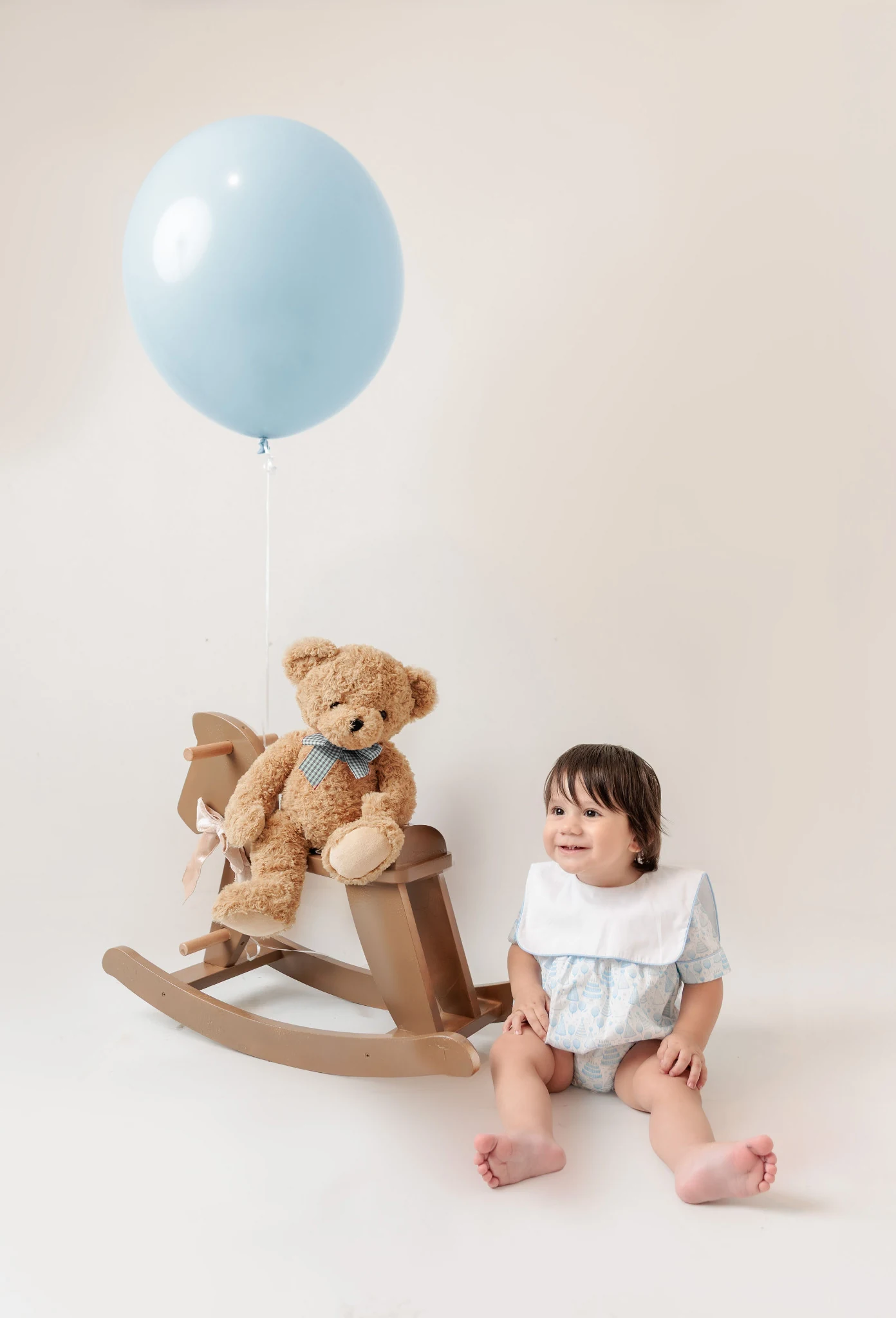 Smiling baby wearing a pale blue patterned outfit beside a teddy and rocking horse
