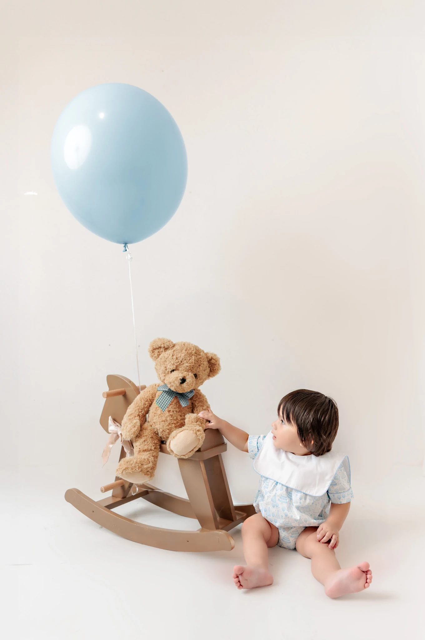 Baby in a pale blue patterned outfit reaching toward a teddy on a rocking horse